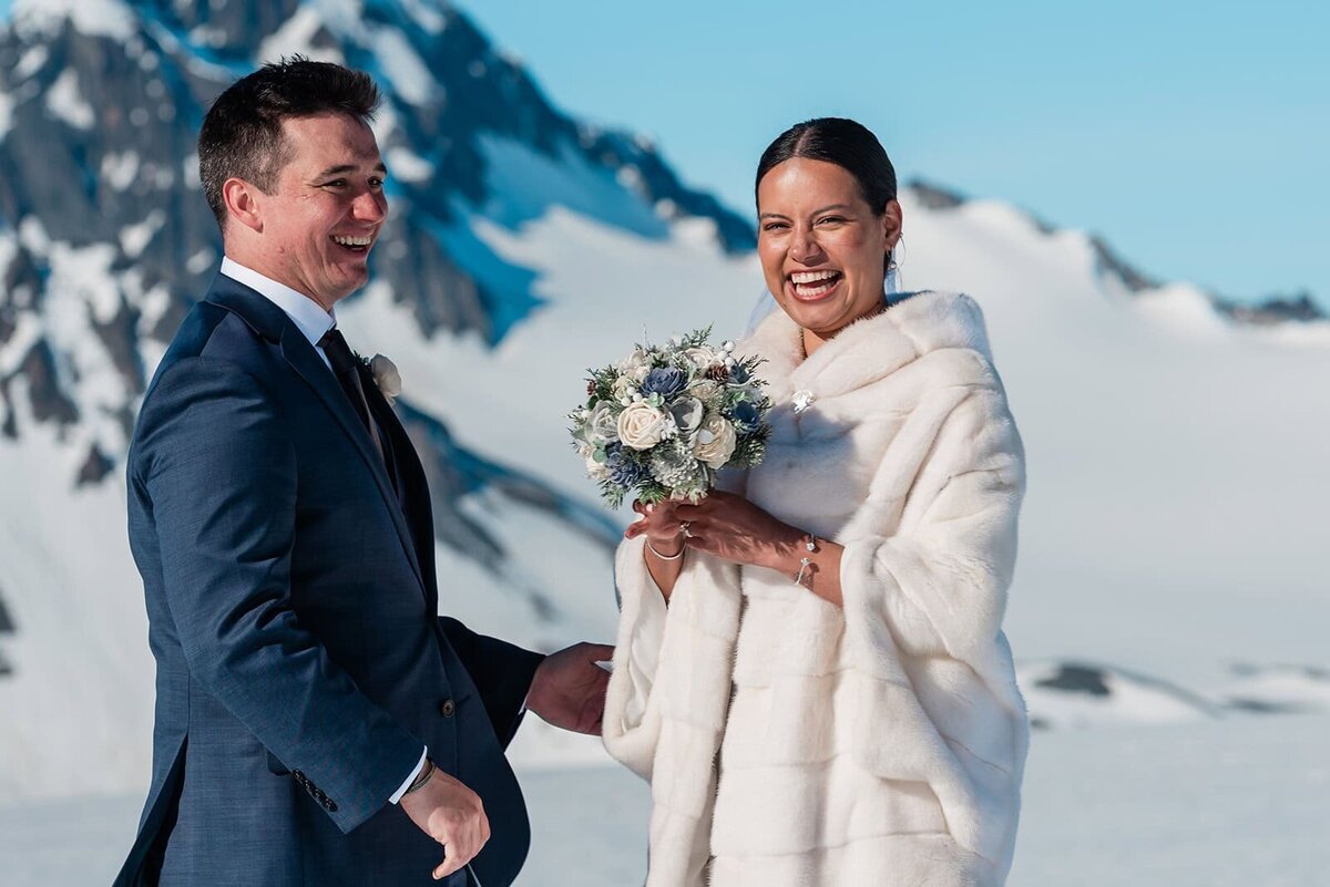 A bride and groom in their wedding finest laugh and smile after just having exchanged elopement vows on the glacier during their two day adventure luxe Alaska elopement.   The sky is bright blue and the snow gleams white against the dark mountains rising up from the glacier.  The bride holds her bouquet and smiles huge to the camera, and the groom is looking to her and smiling a huge smile. 