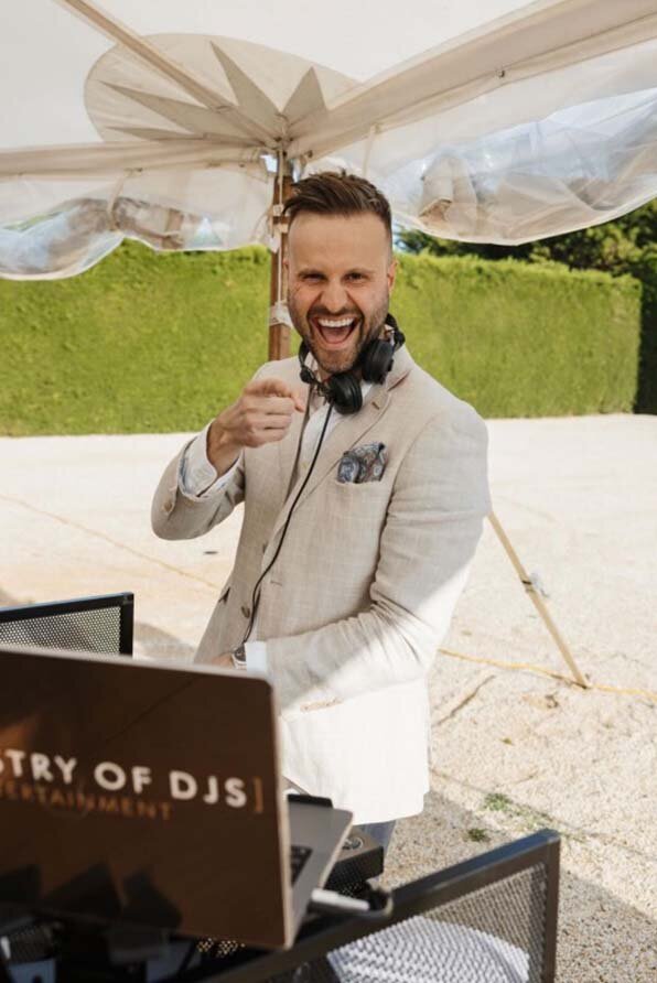 Male DJ in beige suit signalling a thumbs up behind DJ decks at an Australian outdoor wedding event.