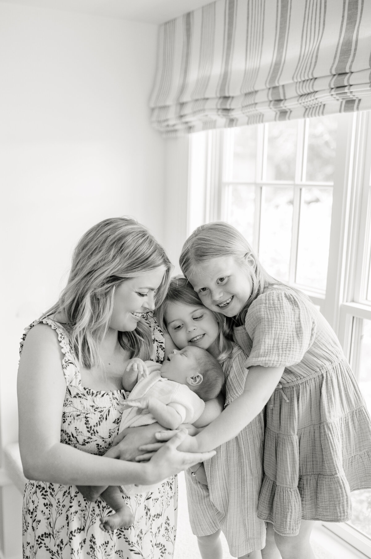 Mom stands holding newborn baby while two older daughters stand on a window ledge to snuggle baby brother