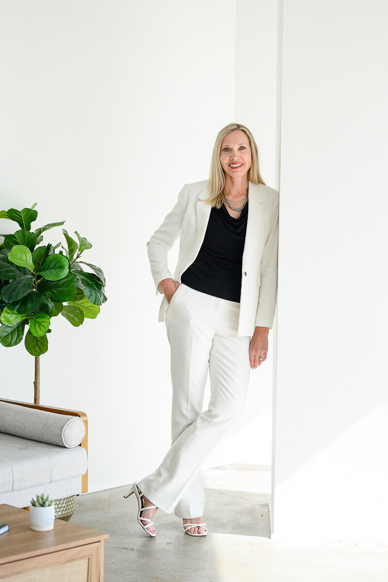 Brand photographer takes a photo of a woman business owner leans on a wall in a white suit taken for her brand photos and her brand photography session.