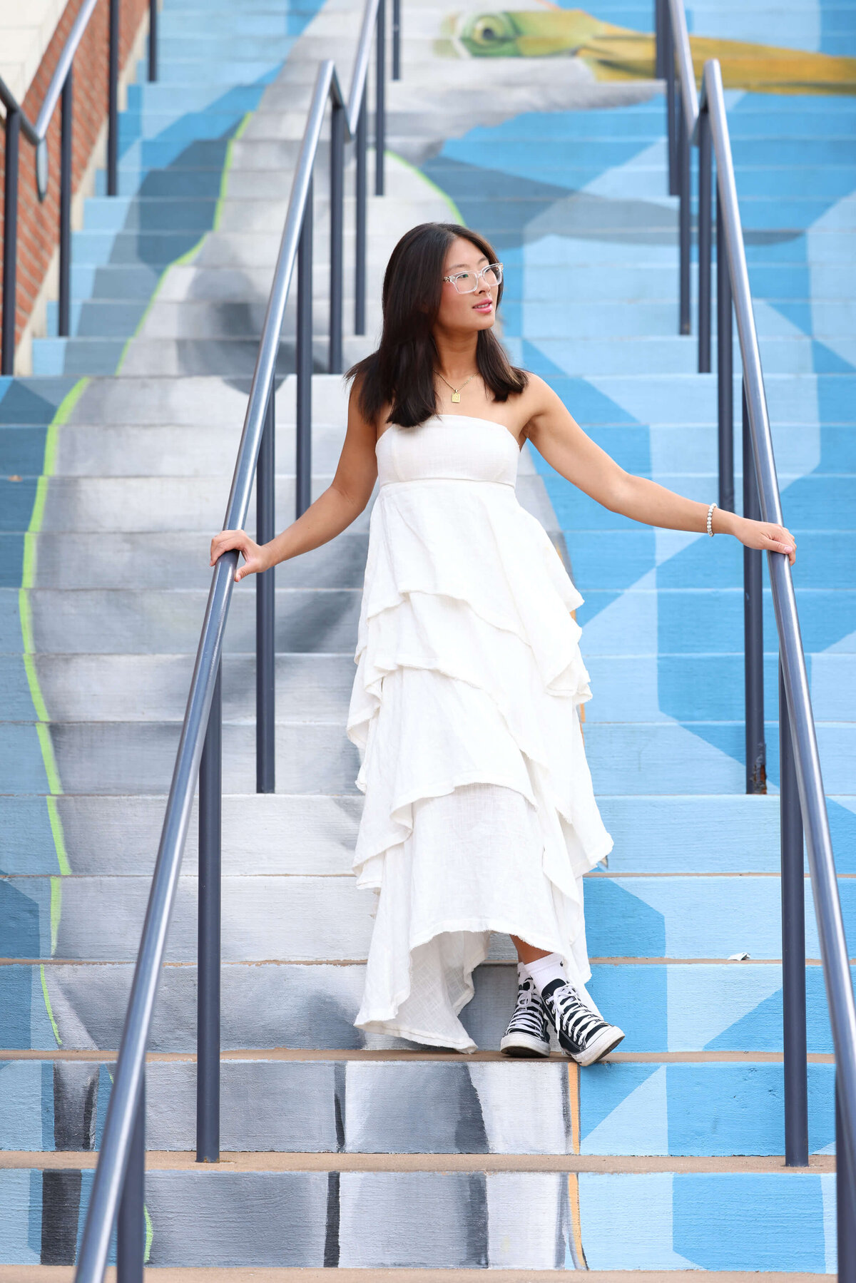 Annapolis High School Senior in flowing white dress and tennis shoes poses on blue heron mural stairs at Annapolis Town Center