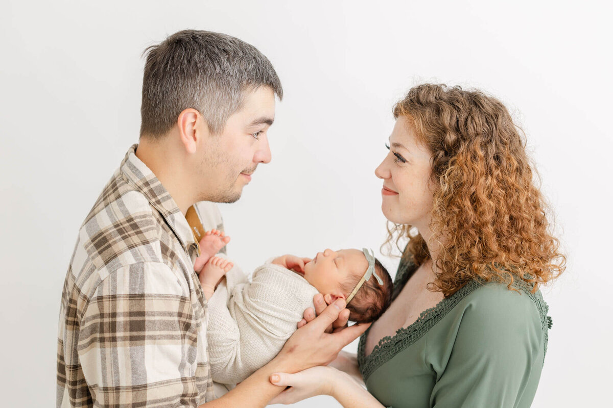 Dad holding newborn baby in front of him while mom stands across from him at baby's head. Mom and dad are looking at each other lovingly and smiling.