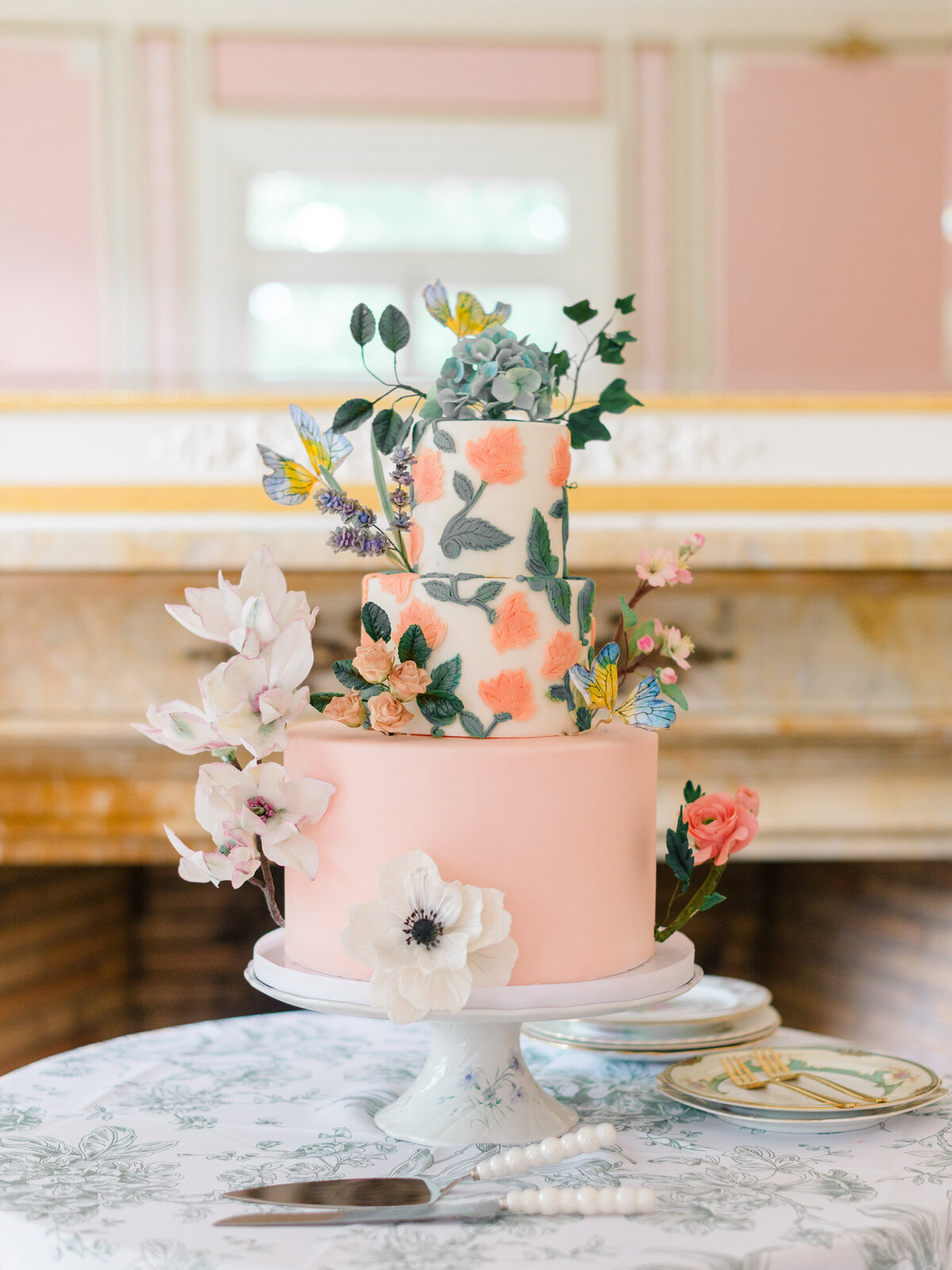 Whimsical three-tiered floral wedding cake with intricate sugar flowers and butterfly details, styled at Cairnwood Estate for a luxury editorial shoot. Captured by Elizabeth Kane Photography, Philadelphia Fine Art Wedding Photographer, this pastel pink statement cake embodies romantic garden elegance and bespoke celebration design.
