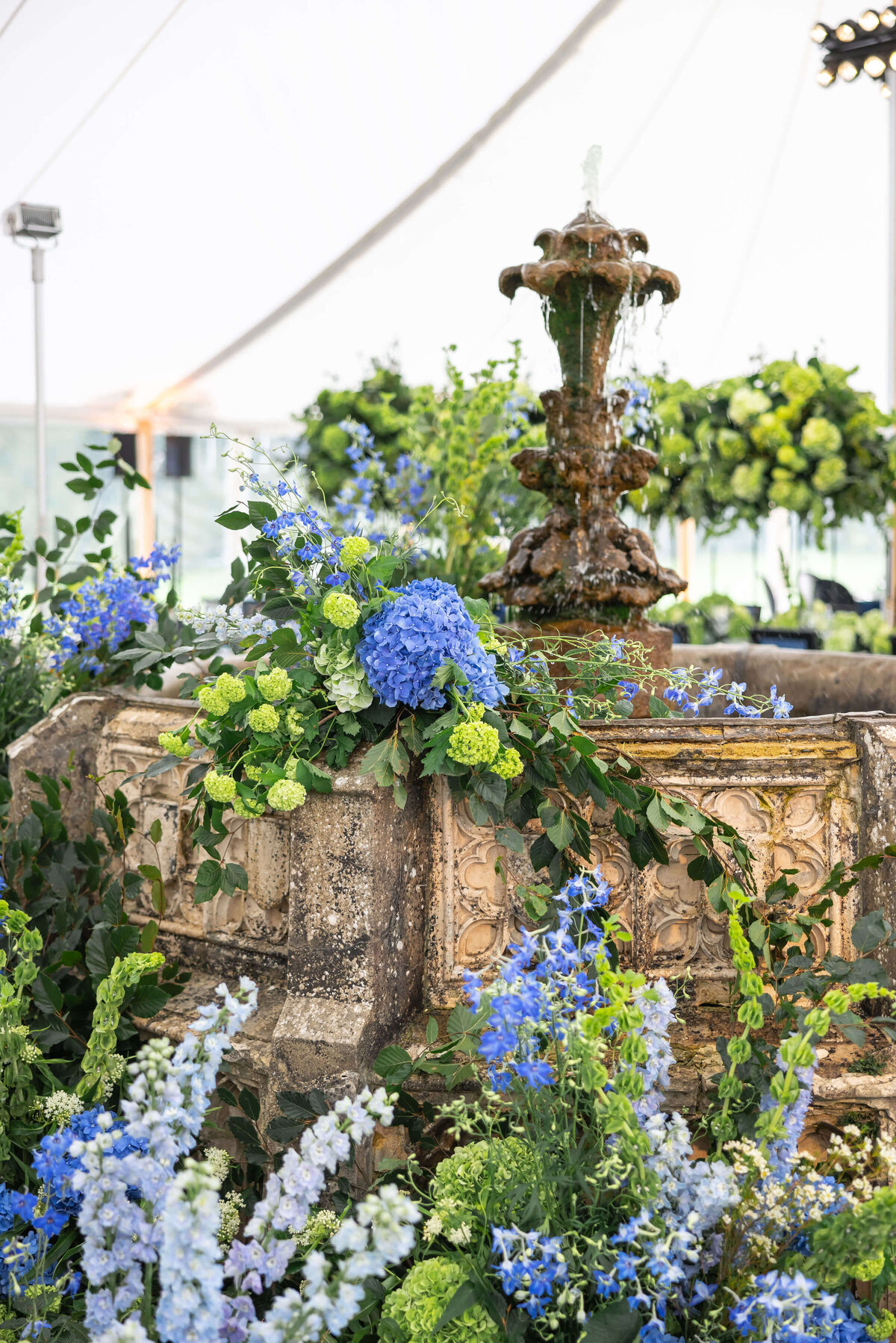 a stone garden fountain decorated with blue and lime green flowers built into the middle of a canvas marquee for a 50th birthday party at avington park
