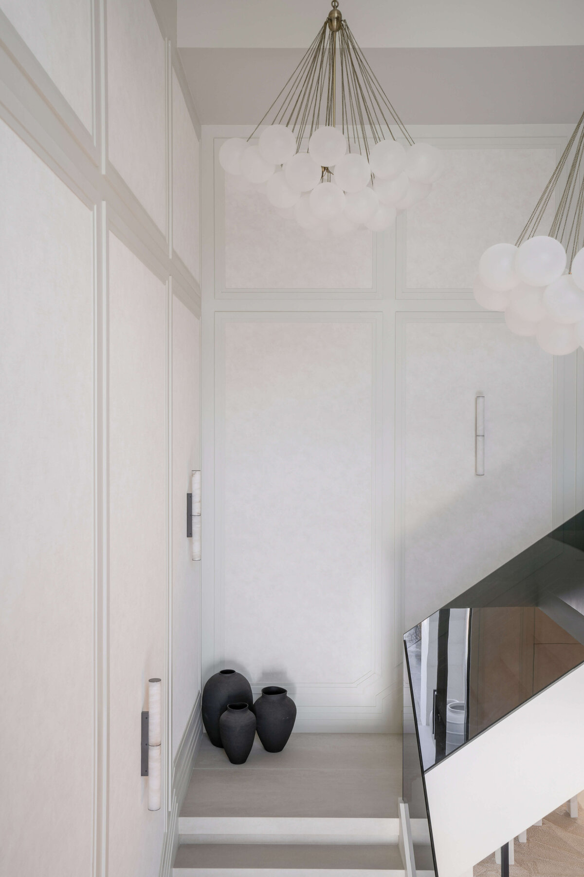 A modern staircase with a minimalist design featuring wide, white steps. The stairs are bordered by a sleek, black glass railing on the right side. At the landing of the staircase, black decorative vases are visible, adding a touch of contrast to the light-colored interior.