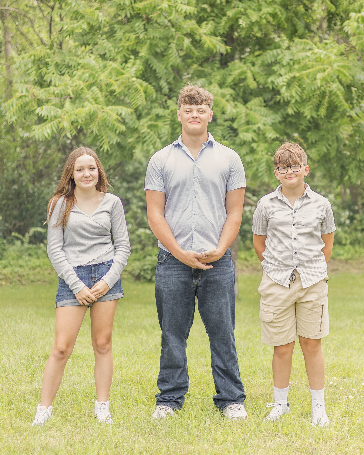 Two brothers and a sister standing next to each in the grass in front of a tree line, smiling at the camera.  Oldest is wearing a light blue button up shirt, blue jeans, white shoes, closed smiling at the camera, and has his arms on his siblings heads giving thumbs up.  Younger brother is wearing a light tan button up shirt, khaki shirt, white shoes, and smiling at the camera.  Sister is wearing a 3 quarter sleeve light blue shirt, jean shorts, white shoes, dark hair, smiling at the camera. 