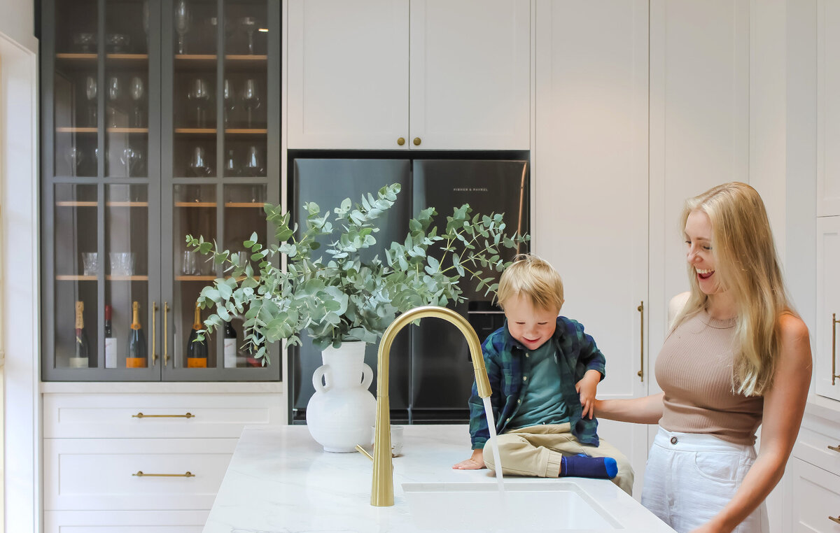 A woman and a child smile in a bright kitchen with white cabinets, designed by a Newcastle Kitchen and Bathroom Designer, while a green plant adorns the counter.