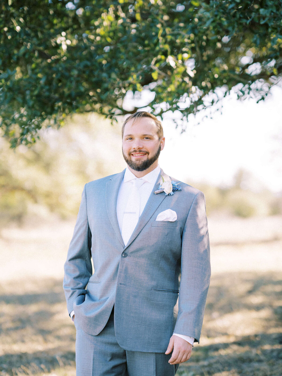 Groom in grey suit with white tie stands under oak tree at The Vineyard at Florence