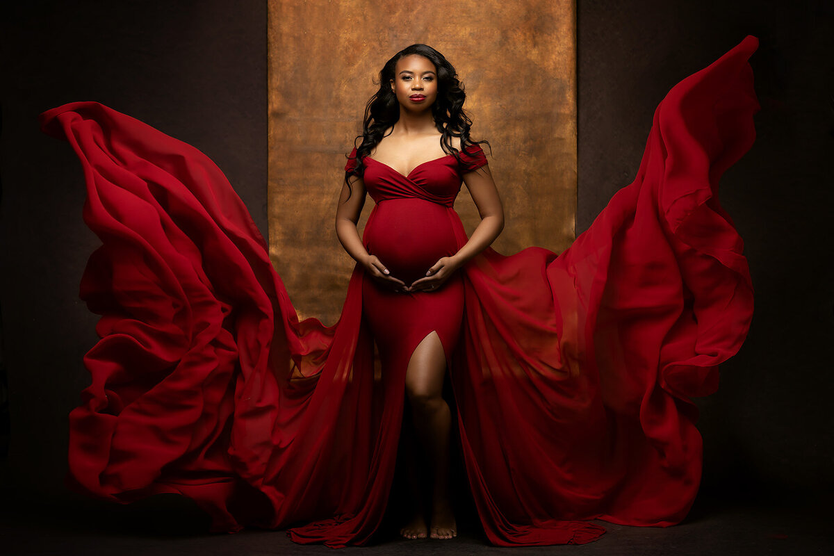 Pregnant woman in a flowing red dress stands barefoot against a dark background, holding her belly with both hands—a serene moment captured perfectly by DFW maternity photography Lindsay Walden.