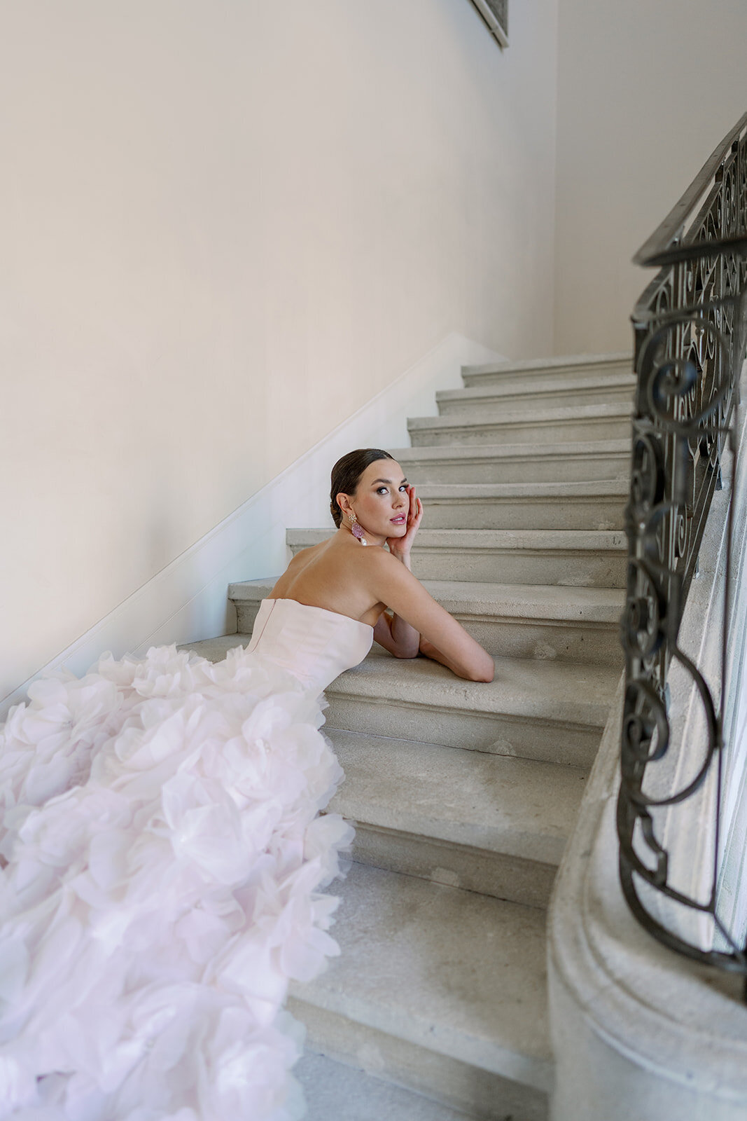 Bride lying on elegant stone staircase showcasing dramatic ruffled blush wedding dress at Château de Tourreau.