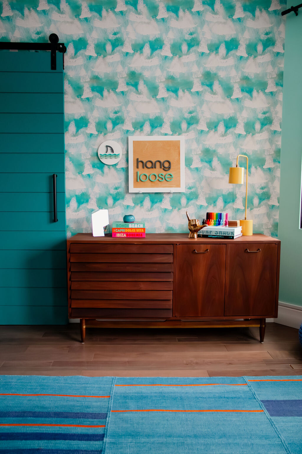 A mid-century modern wooden dresser stands against a wall with a vibrant turquoise and white patterned wallpaper. On the dresser, several stacks of colorful books are arranged. On the left, a stack includes books titled "BONDI BEACH," "CAPRI DOLCE VITA," and "IBIZA BOHEMIA." A small, translucent object and a teal glass ball sit atop this stack. In the center, a shiny gold-colored hand sculpture making the "hang loose" shaka sign rests on a stack featuring a book titled "SURF SHACK." A set of colorful markers is also placed on this stack. A small framed print with the words "hang loose" in teal lettering on a wood-look background hangs on the wall above the dresser, next to a round clock with a shark fin. A slender gold-toned desk lamp with a cylindrical shade stands on the right side of the dresser. To the left, a teal-painted wooden door with a long black handle is partially visible.
