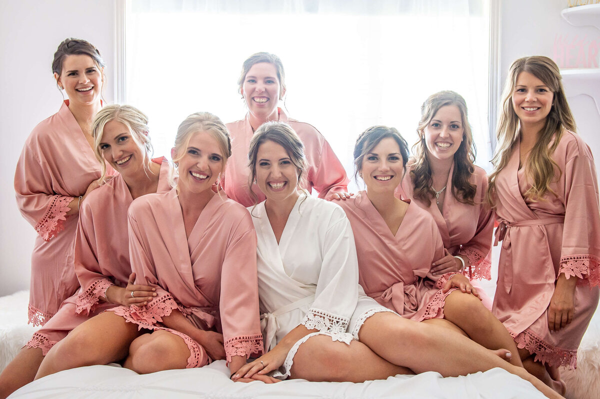 A bride sitting on her bed surrounded by her bridesmaids in pretty pink robes as she gets ready for her Ottawa wedding