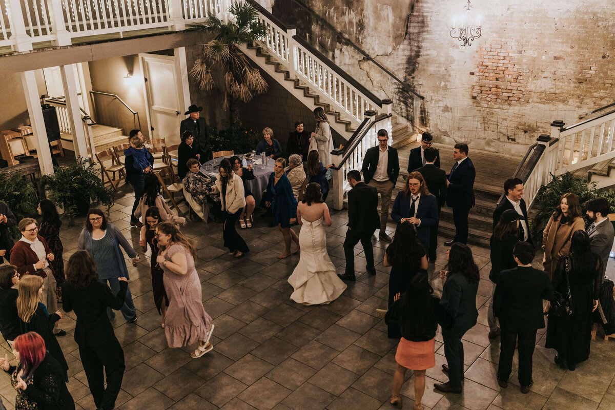 A group of people in formal attire gather and mingle on a tiled floor at an outdoor venue with stairs, plants, and exposed brick walls. A bride in a white dress stands at the center of the scene.
