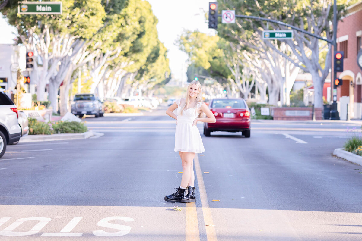 A woman in a white dress and black boots stands confidently in the middle of a street. Trees line the road, and traffic signals are visible above. A red car approaches from behind. Its a sunny day, and the scene conveys a relaxed, urban vibe.