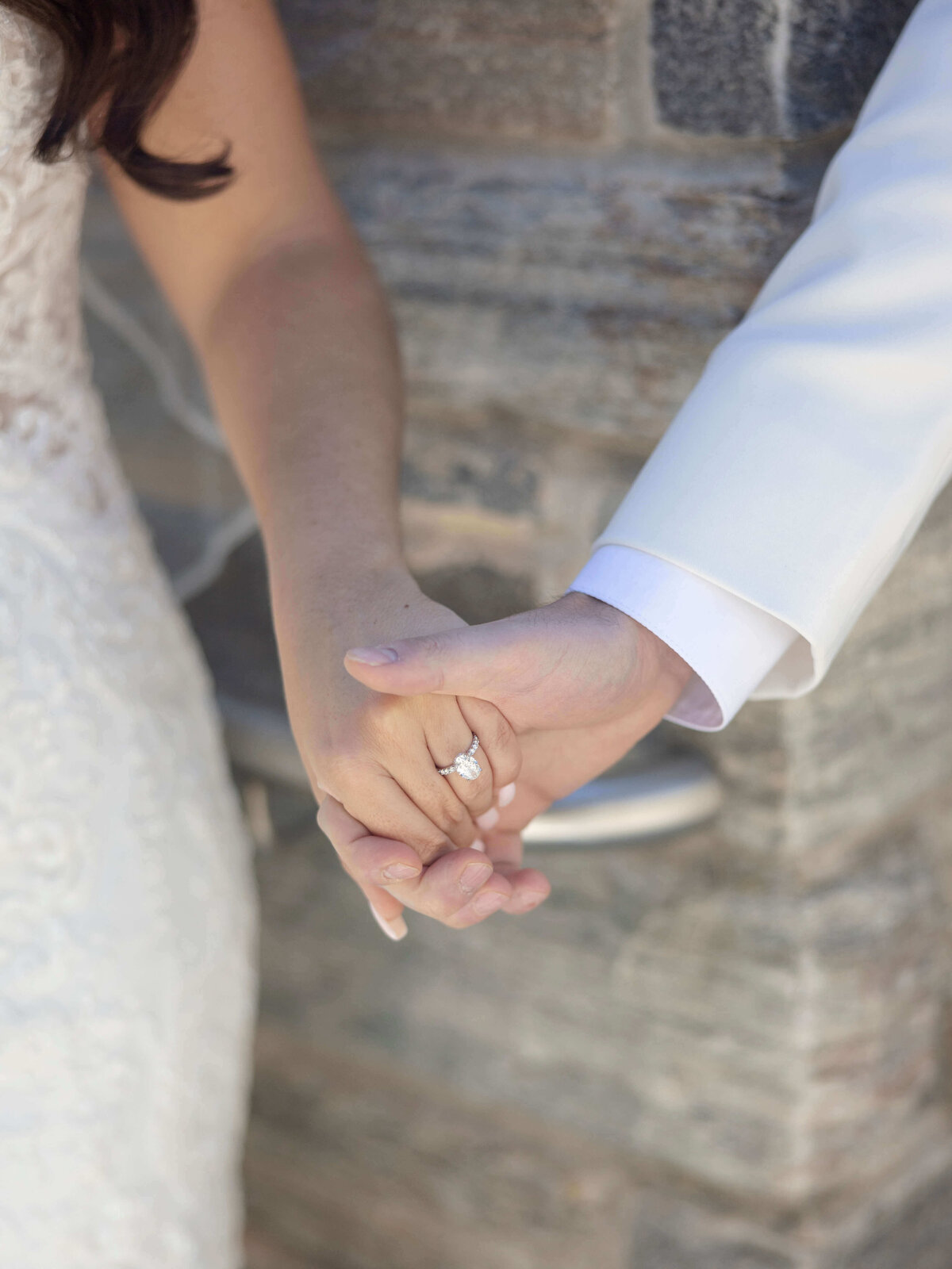 Close-up of a couple holding hands, showcasing an engagement ring on one hand. The person on the left is wearing a white lace dress, and the person on the right is wearing a white suit. They are standing in front of a stone wall.