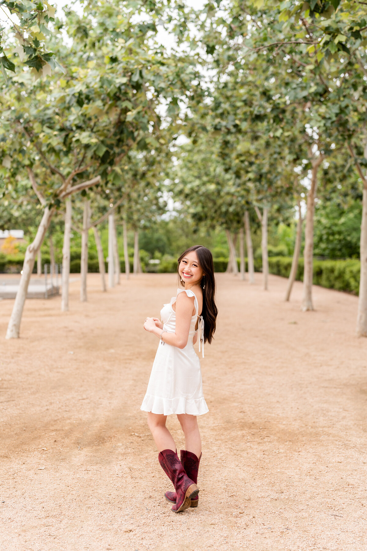 High School senior girl looking back over shoulder while wearing white dress and maroon boots in middle of park in Downtown Houston