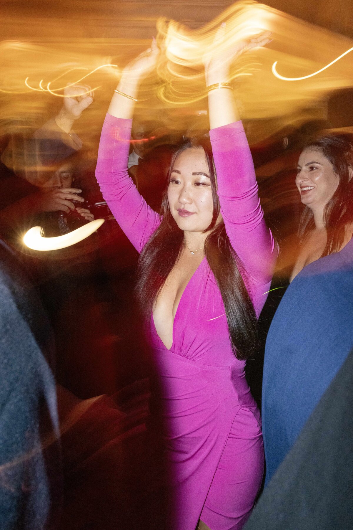 A woman in a vibrant pink dress dances with her arms raised amid a dynamic blur of lights. She stands amongst a crowd, exuding joy and energy. Another person in a blue outfit smiles nearby, capturing the lively atmosphere of the scene.