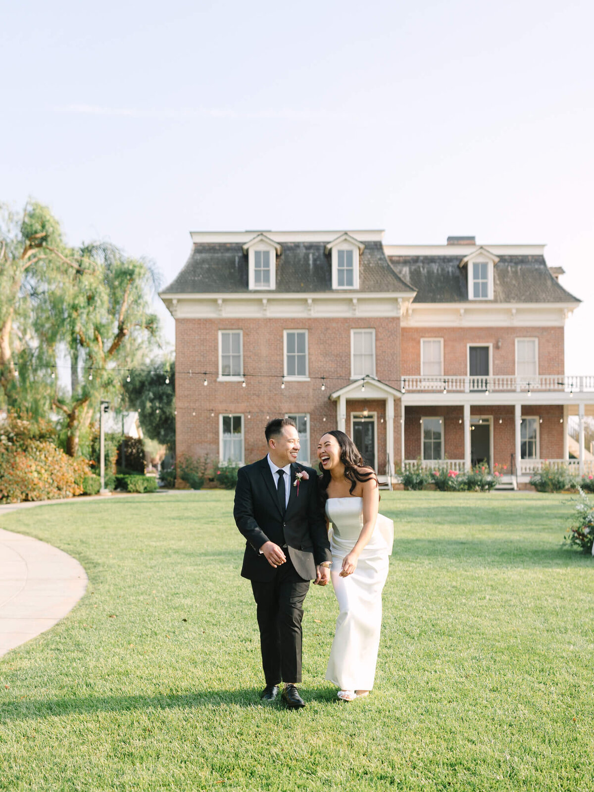 A joyful couple in formal attire walks hand-in-hand on a lush lawn in front of the Barton House.