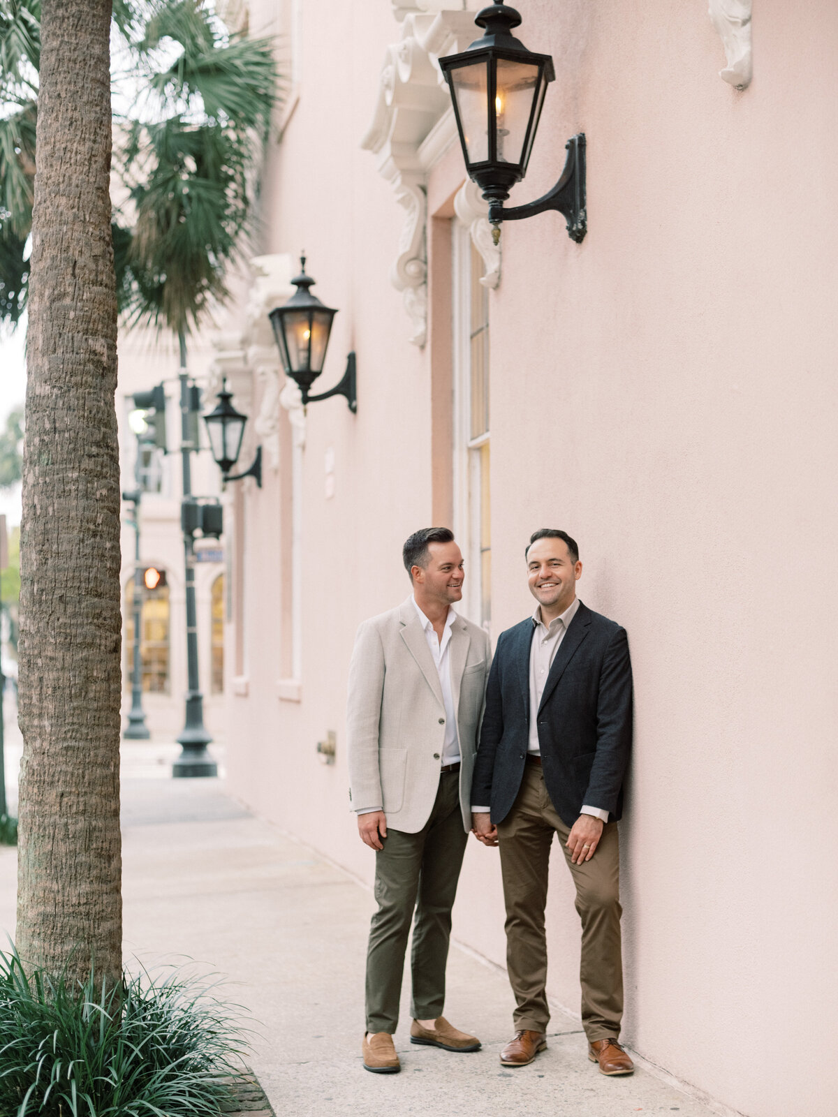 A joyful couple sharing laughter and affection standing together against a charming pink wall in Charleston, capturing the essence of love, companionship, and happiness in life.