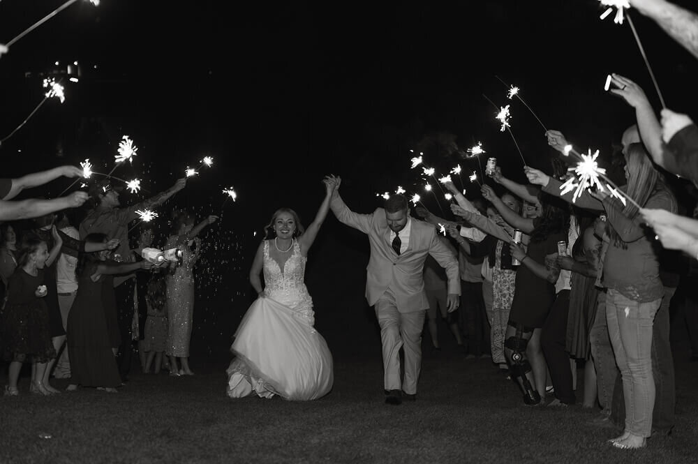 A black and white photo of a bride and groom running through an aisle of people, all holding sparklers.