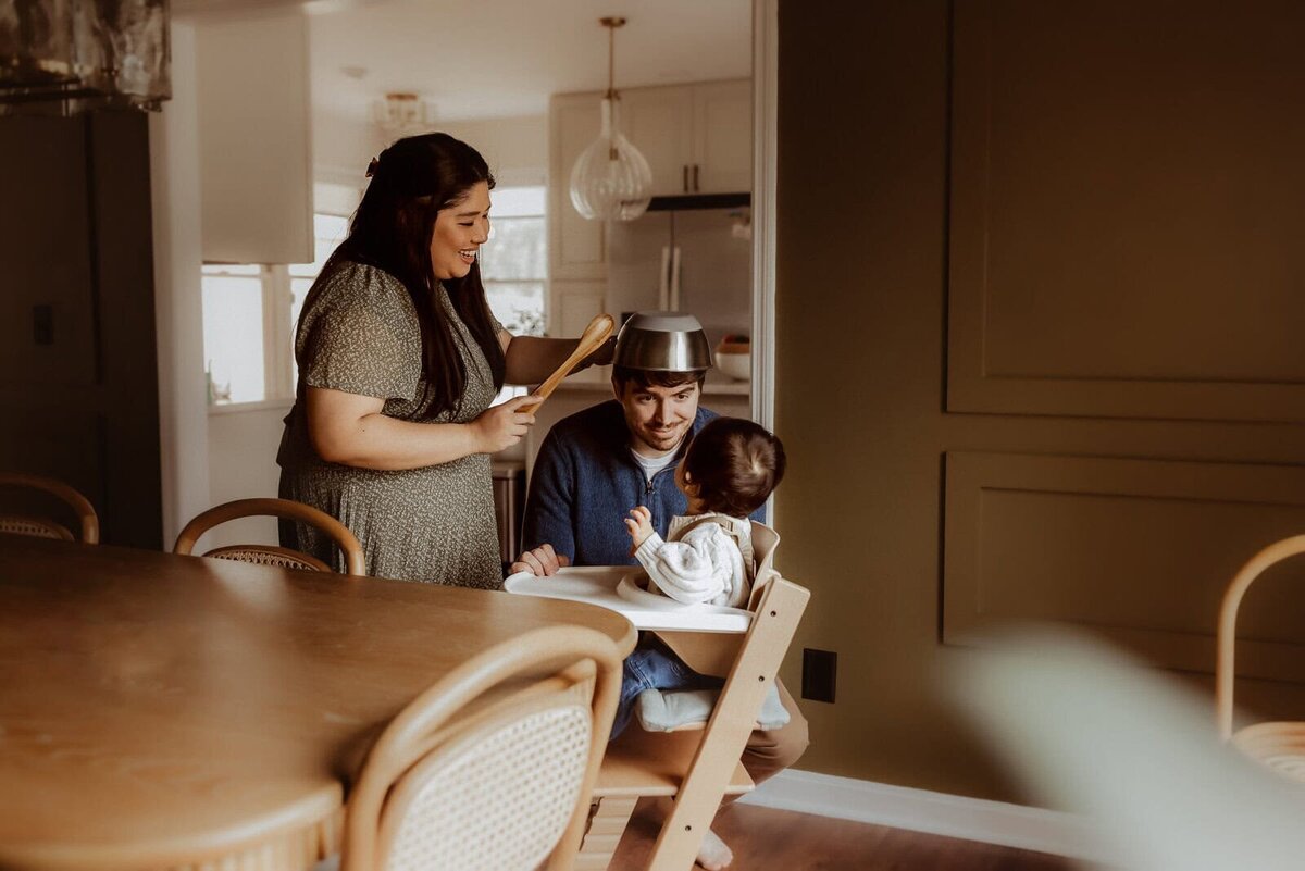 A woman places a pot on a man's head, who is sitting with a child in a high chair. They are all in a dining area with wooden furniture.