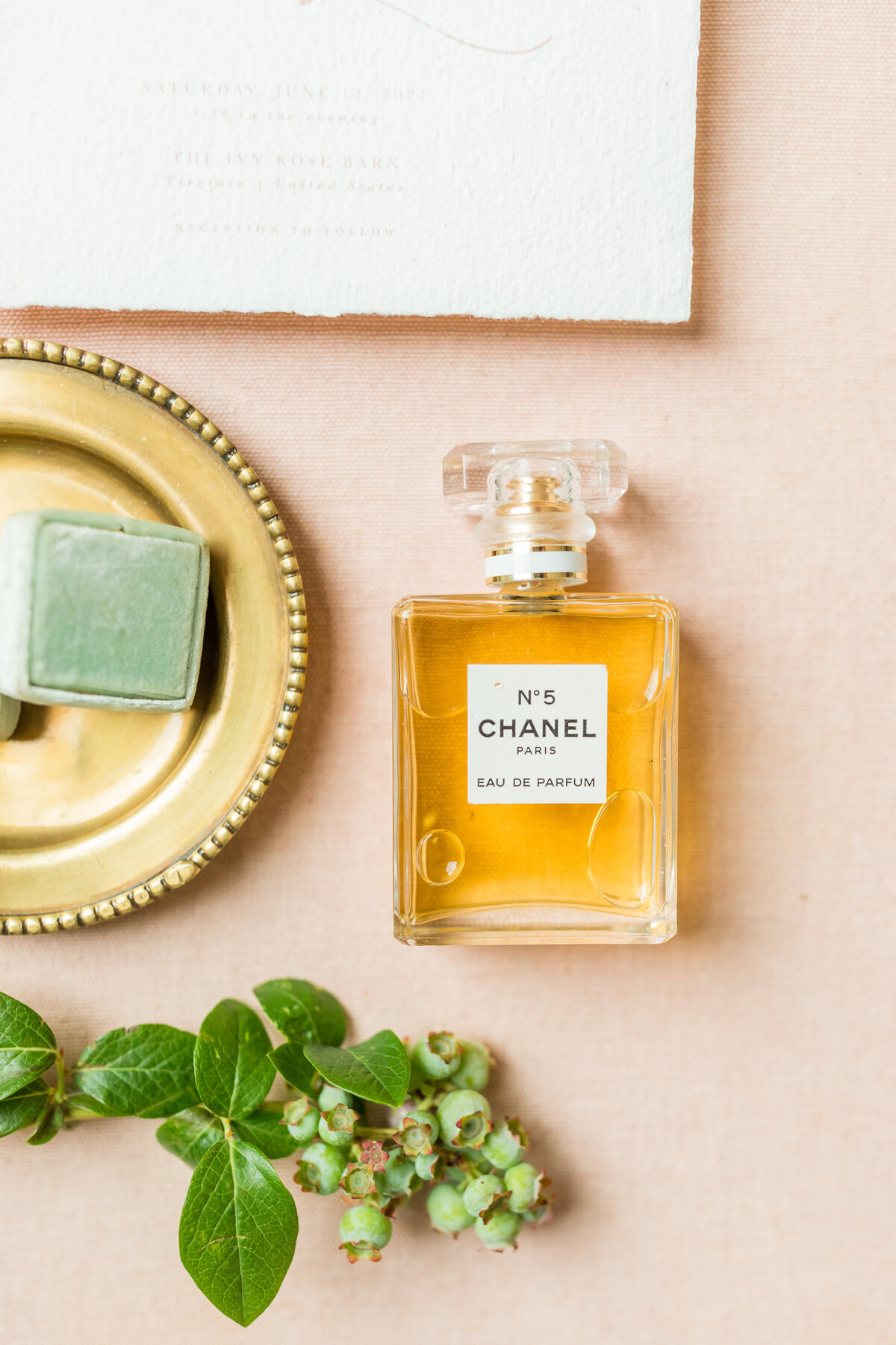A bottle of Chanel No. 5 perfume elegantly styled on a soft pink surface, accompanied by a gold tray, a green ring box, and delicate greenery for a refined and timeless aesthetic.