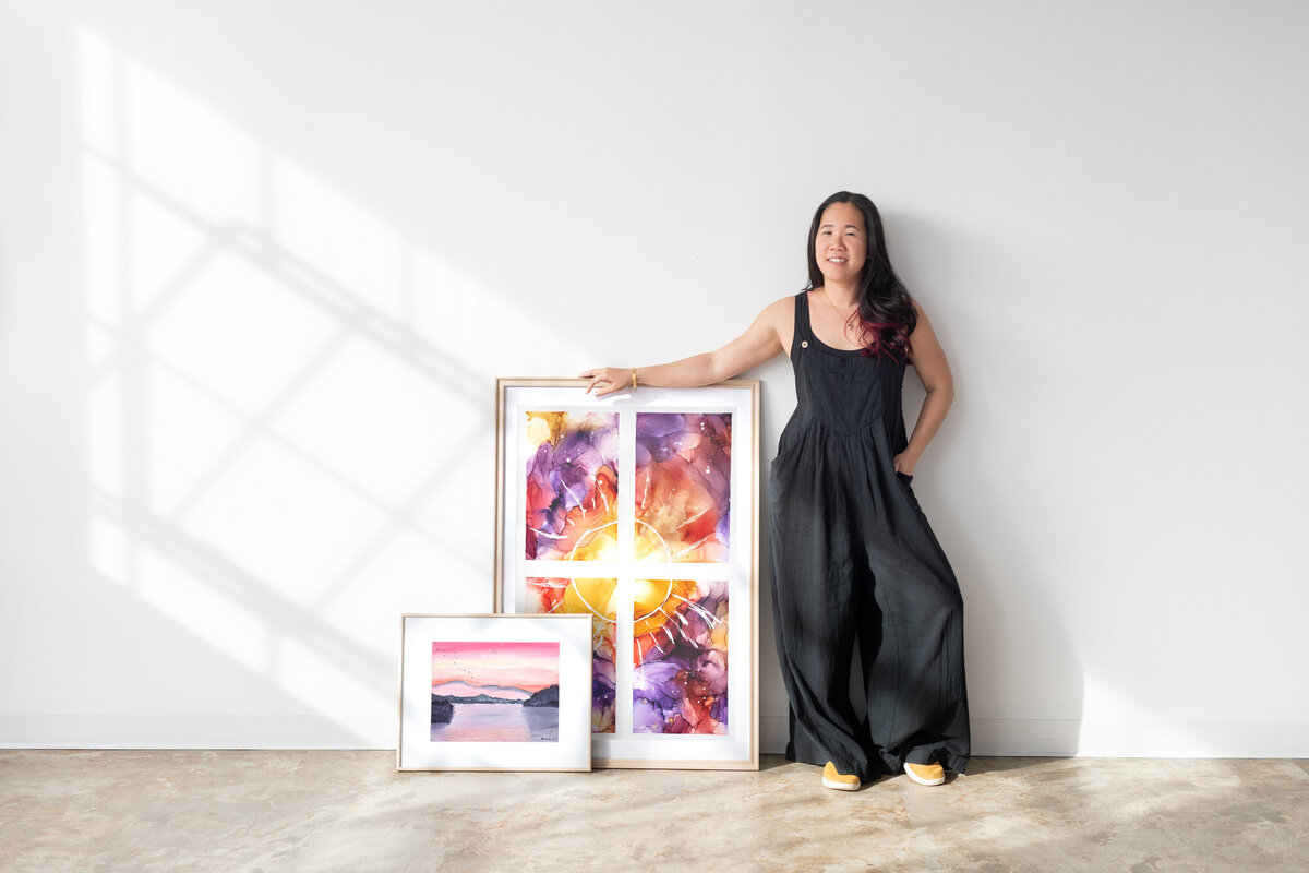 asian woman wearing yellow loungers and black overalls standing with her framed artwork
