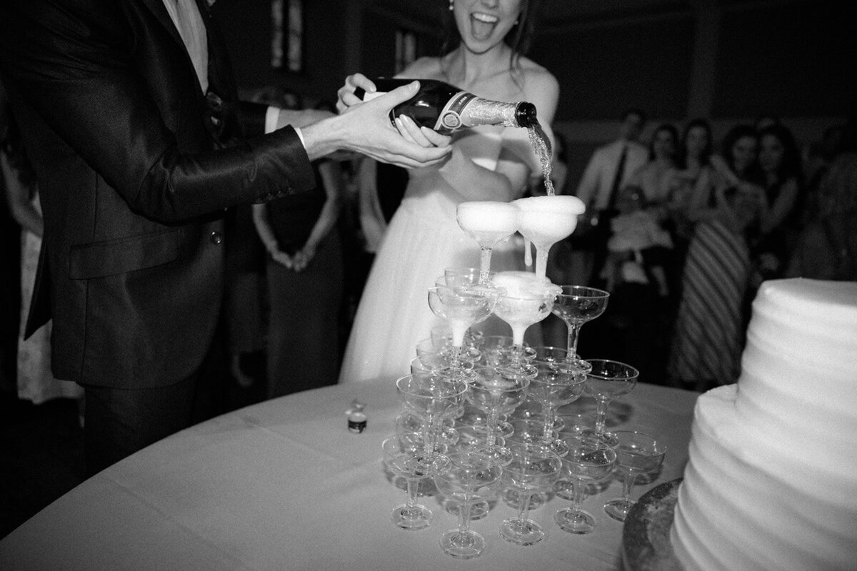 Bride and groom pour champagne into a champagne tower during their wedding reception festivities while laughing. 
