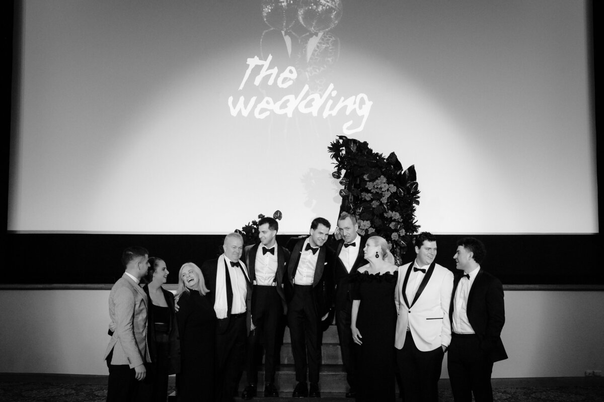 The grooms at The Wedding in Regal Cinema with their families all in front of the stage.