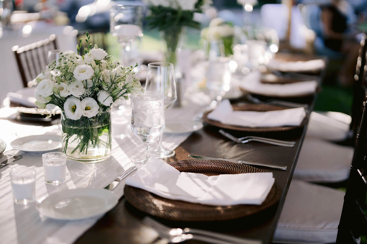 A dining table with a white table runner, white flower centerpiece, cutleries, and wine glasses in Cape Cod Summer Tent, MA