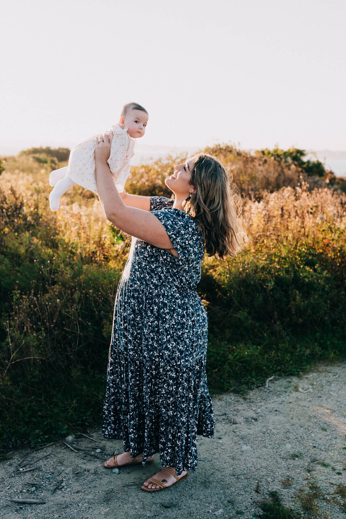 A mother wearing a blue and white floral dress holds up and looks at her baby girl in a field at sunset