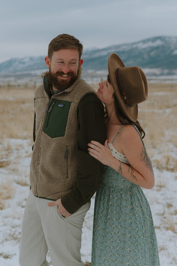 A wife holds her husband's arm from behind as she looks up at him and smiles. He's looking back at her too.
