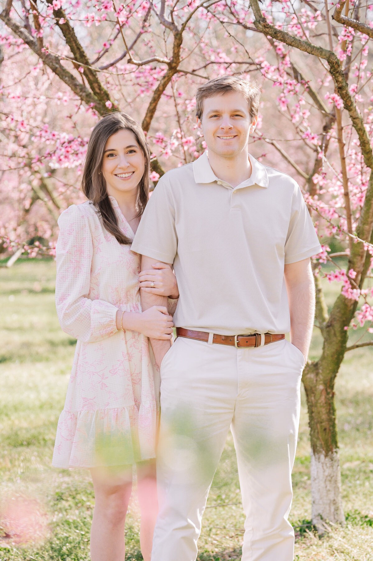 A smiling engaged couple standing in bright pink cherry blossoms in the North Carolina countryside at sunset enjoying their destination wedding photos by JoLynn Photography