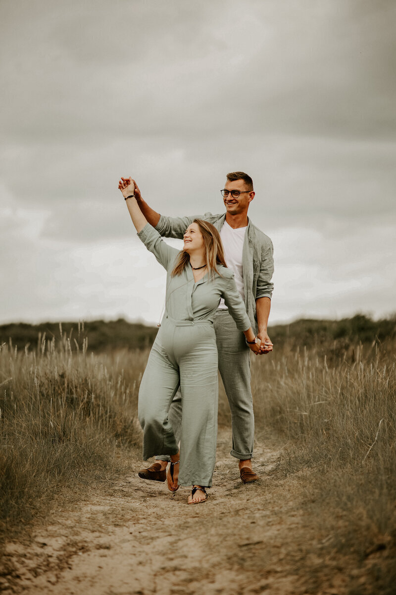 Femme enceinte marchant dans les dunes. Derrière elle son chéri qui lui tient les mains. Séance photo grossesse en vendée.