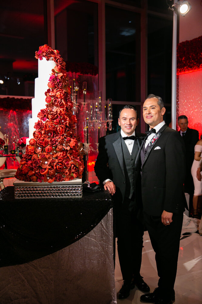 Two grooms standing next to a tall wedding cake covered in red roses