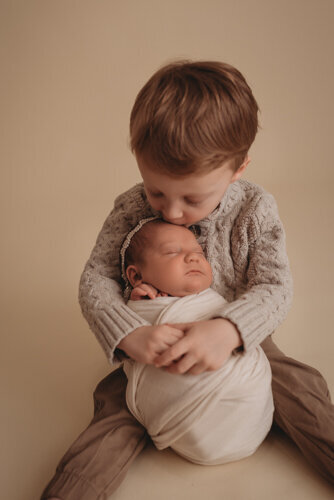 Baby girl sleeping for newborn portrait session with Atlanta Georgia newborn photographer snuggled up in a swaddle posing with her big brother giving her a kiss on her forehead