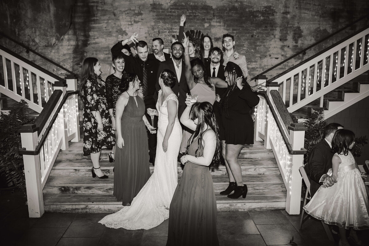 A bride and groom stand on stairs surrounded by joyful friends and family, some posing, smiling, or laughing. The group is dressed formally. The setting appears festive, with string lights on the railings. The mood is celebratory.