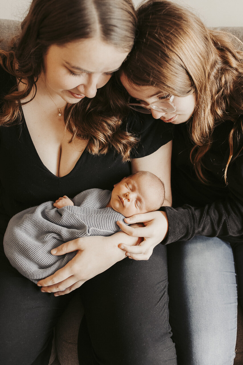Mom and sister looking down at precious newborn wrapped in blue blanket, sleeping: lifestyle newborn photography in Tifton Georgia