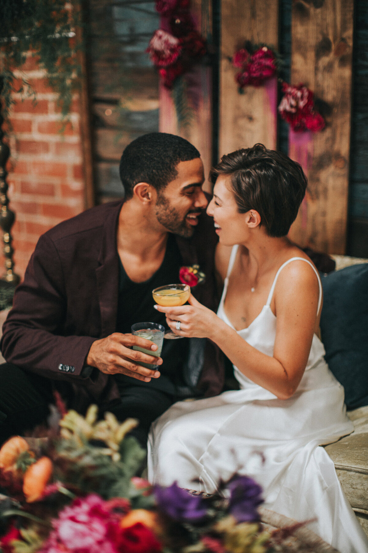Bride and groom toast to their wedding while nuzzling on a sofa surrounded by flowers