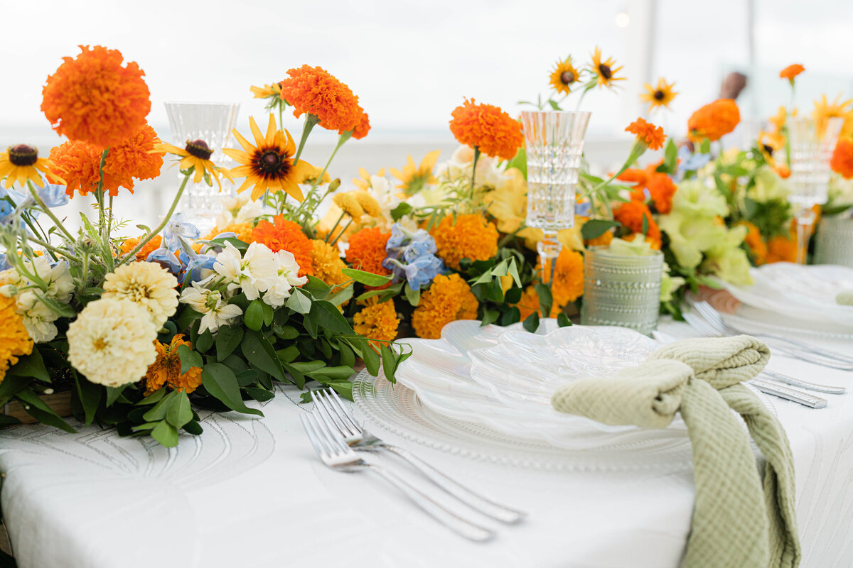 white on white palm leaf tablecloth with hammered silver forks and an iridescent seashell plate adorned with a white, orange, blue and green floral table runner and green gauze napkin tied in a bow