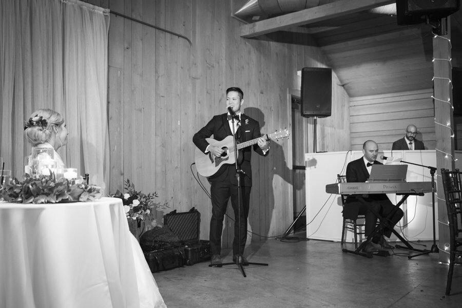 A groom plays acoustic guitar as he serenades his bride at their wedding reception.