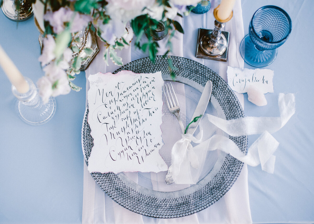 Beautiful wedding calligraphy cards sit atop a silver charger  plate with cutlery on luxury wedding table.