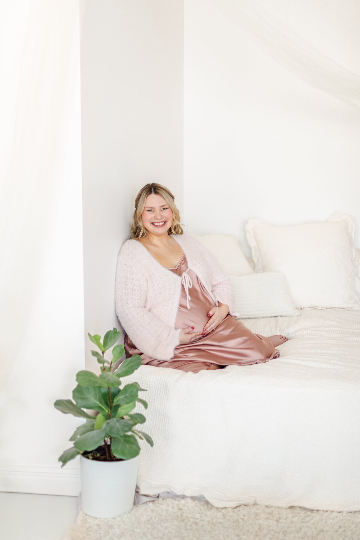 A peaceful and elegant image of a mother sitting on a bed, wearing a flowing silk dress, lovingly holding her baby bump. A delicate plant rests beside the bed, adding a soft, natural touch.