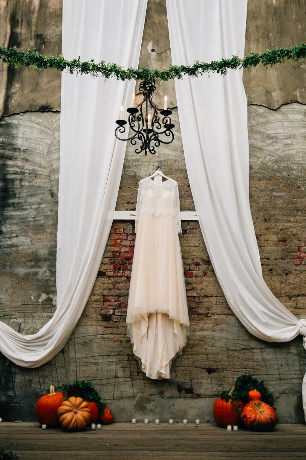 A wedding dress hangs against a brick wall, framed by white drapery. Pumpkins and candles decorate the wooden landing in front of the wall. A black chandelier hangs on green garland above.
