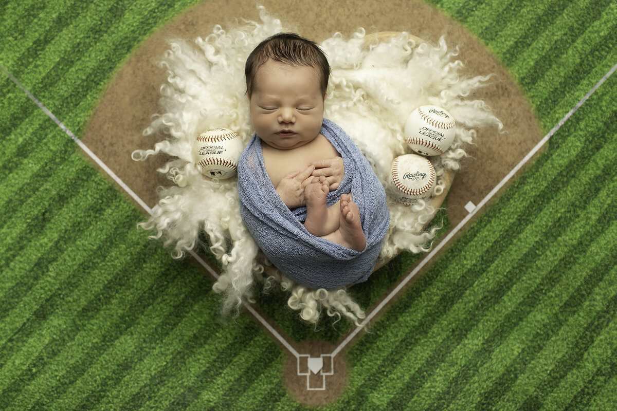 Newborn baby boy snugly wrapped and peacefully resting on a baseball-themed backdrop, creating a sweet and sporty scene