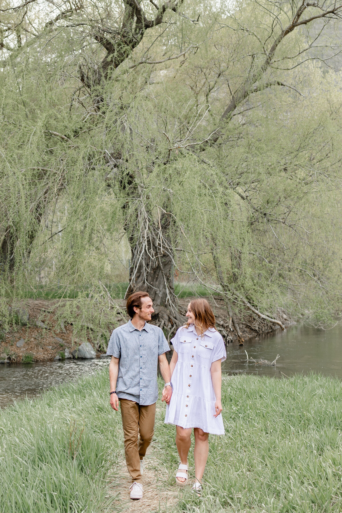 A couple walks hand in hand along a riverside path, smiling at each other.. Take by Cassie Lee Photography