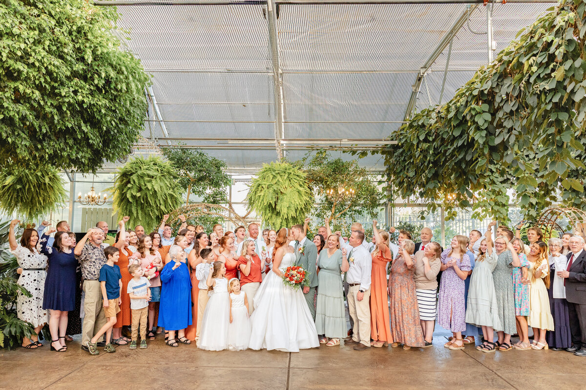 A joyful group portrait of the newlyweds with their family and friends, gathered in Le Jardin’s greenhouse under vibrant hanging greenery