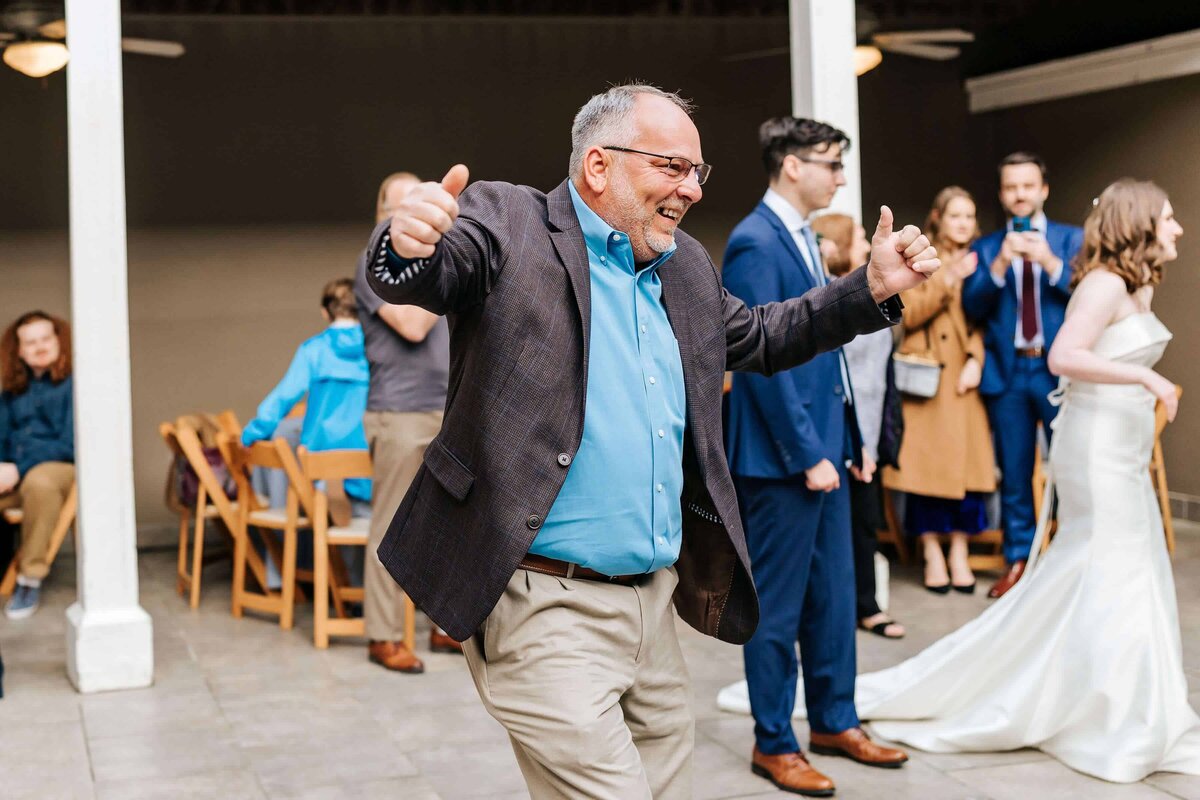 A smiling man in a blazer and blue shirt dances with his thumbs up at a wedding reception, while other guests, including a bride in a white dress, socialize in the background.