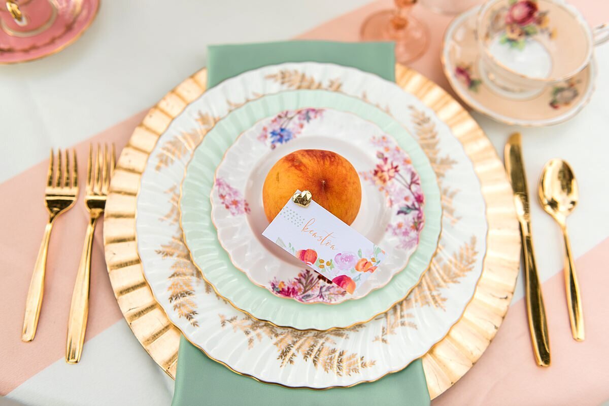 A detail of a place setting at a summer wedding at Ravenswood Mansion. The fluted gold charger and sage green napkin are accented by a vintage china dinner plate, vintage china salad plate and vintage china dessert plate with a fresh peach in the center. A seating card is pinned to the peach. The seating card features hand written modern calligraphy. On either side of the place setting is gold flatware and a vintage china teacup.