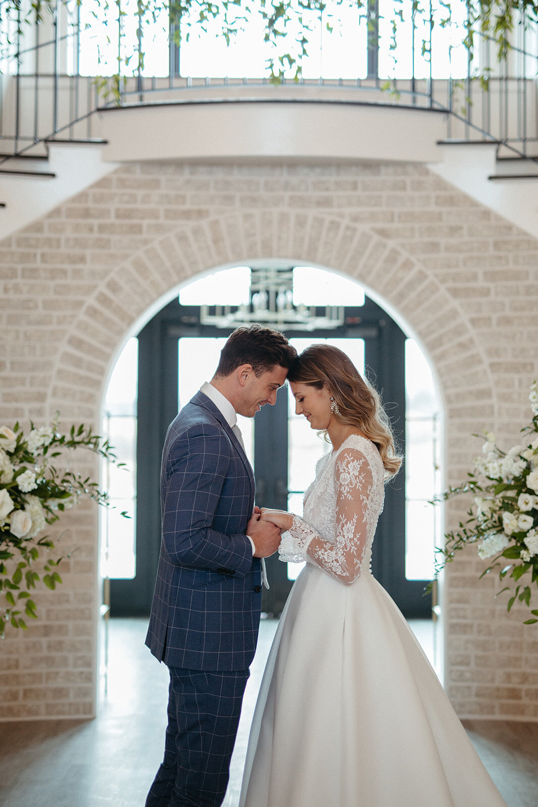 Bride and groom in a blue suit and white wedding gown holding hands in front of a whitewashed brick wall with flowers.